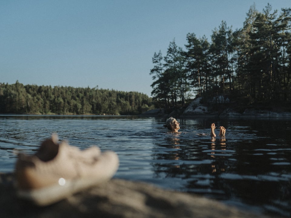 Fotograf: Plattform
Bildbyrå: Johnér Kvinna badar i sjö. Skog i bakgrunden och ett par skor i förgrunden.