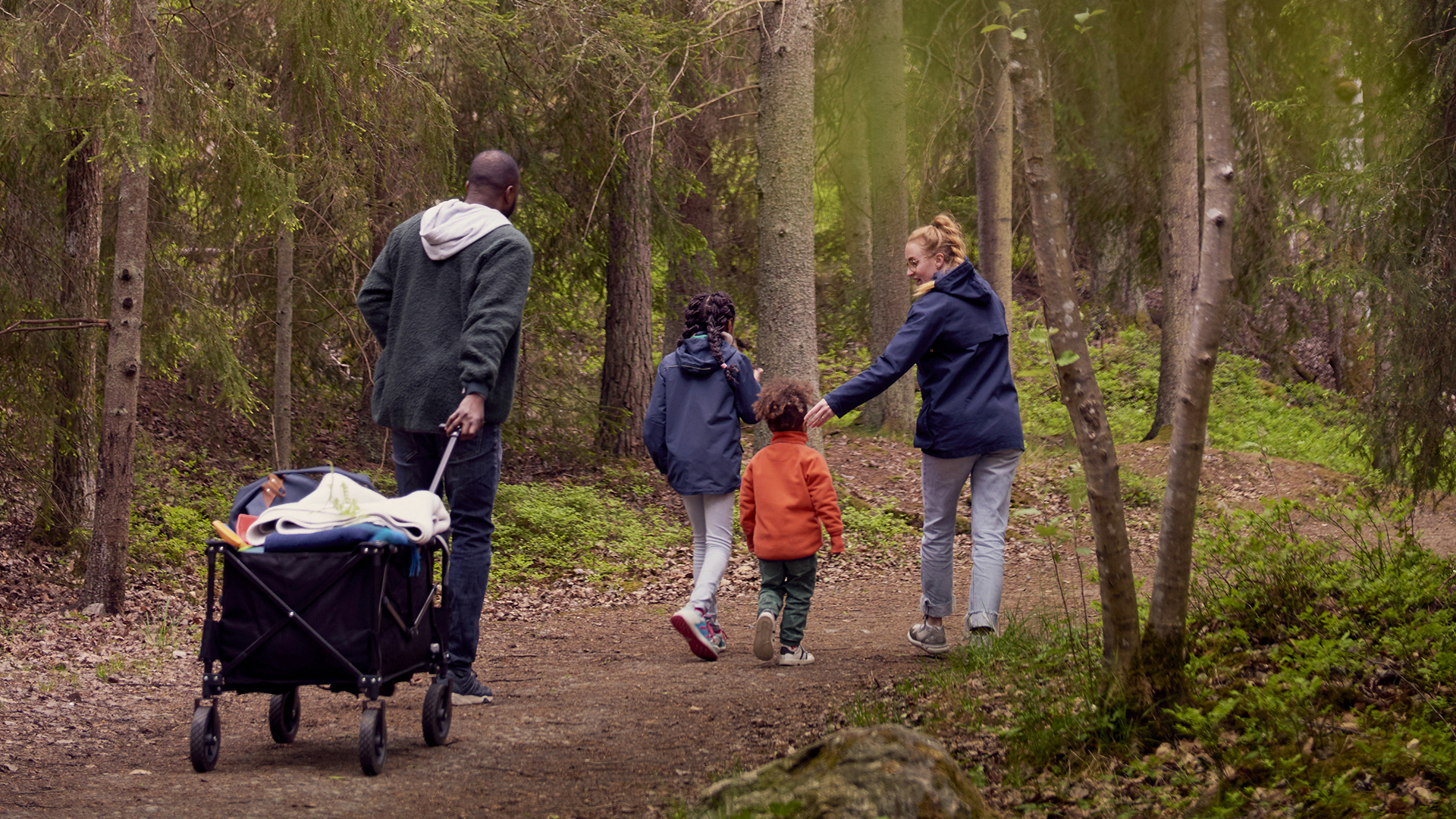 Familj med två barn promenerar i skogen och drar en campingvagn efter sig. Sverige.