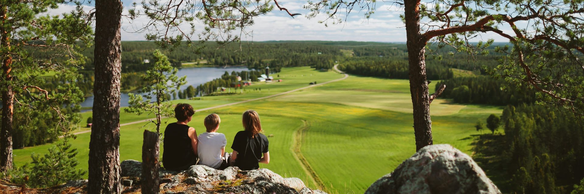 Photographer: Matilda Holmqvist
Agency: Johnér Three people sit on a hill (Falkberget) and look out over a cultivated landscape with a lake.