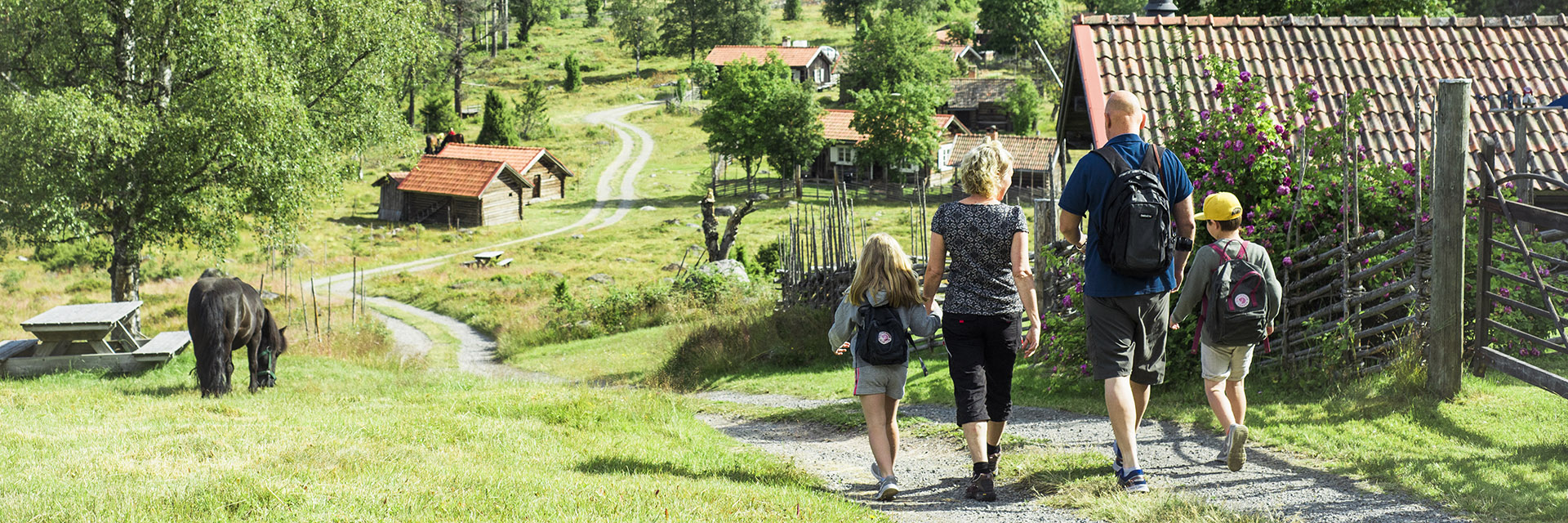 Två vuxna och två barn går på grusväg i en by omringad av träd.