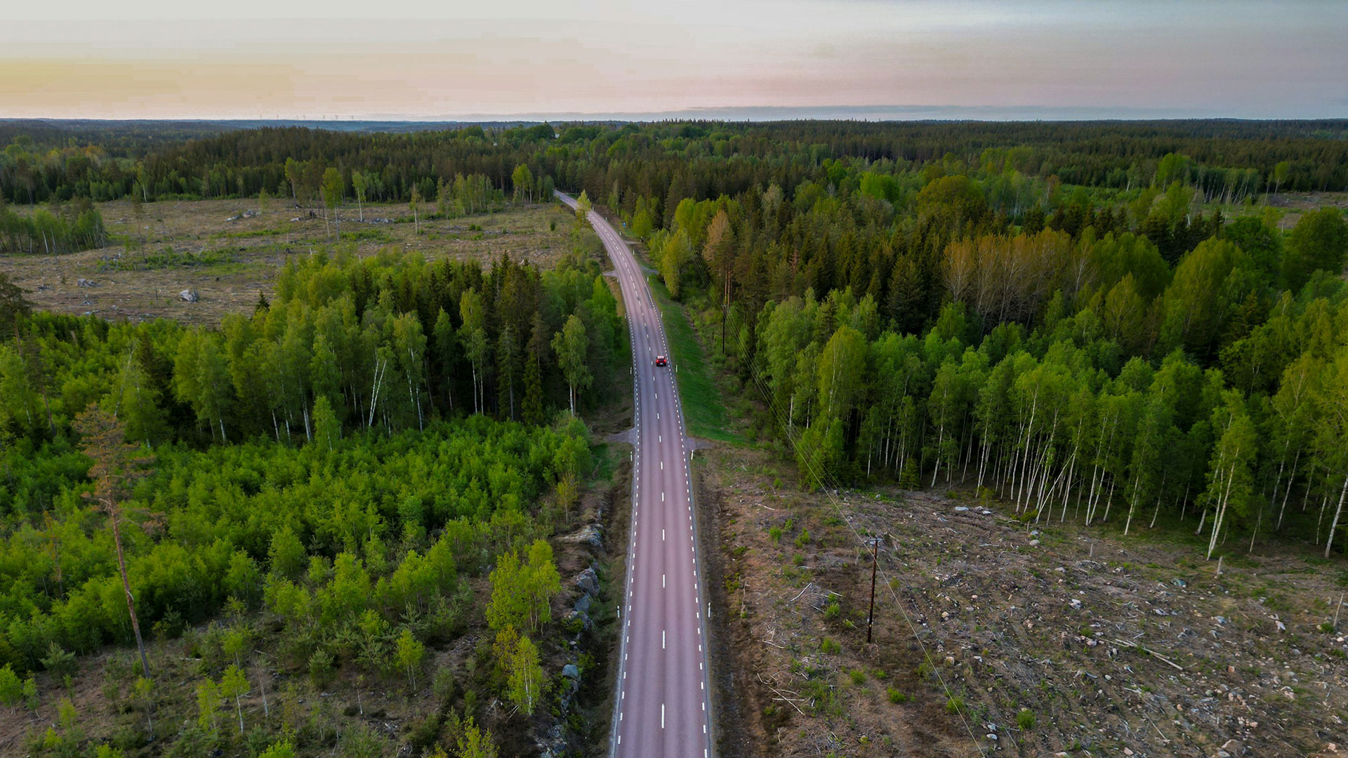 Flygbild av en rak landsväg som skär genom tät grön skog, med ett kalhygge som tydlig kontrast i landskapet.