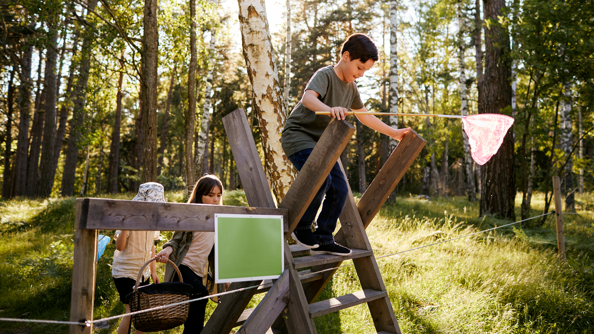 Fotograf: Plattform
Bildbyrå: Johnér Barn på utflykt i skogen med fjärilshåvar och picknickkorg. De klättrar över ett stängsel vid en stätta (en form av trappa över stängslet).