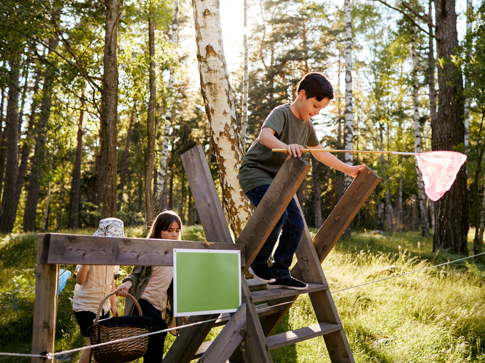 Fotograf: Plattform
Bildbyrå: Johnér Barn på utflykt i skogen med fjärilshåvar och picknickkorg. De klättrar över ett stängsel vid en stätta (en form av trappa över stängslet).