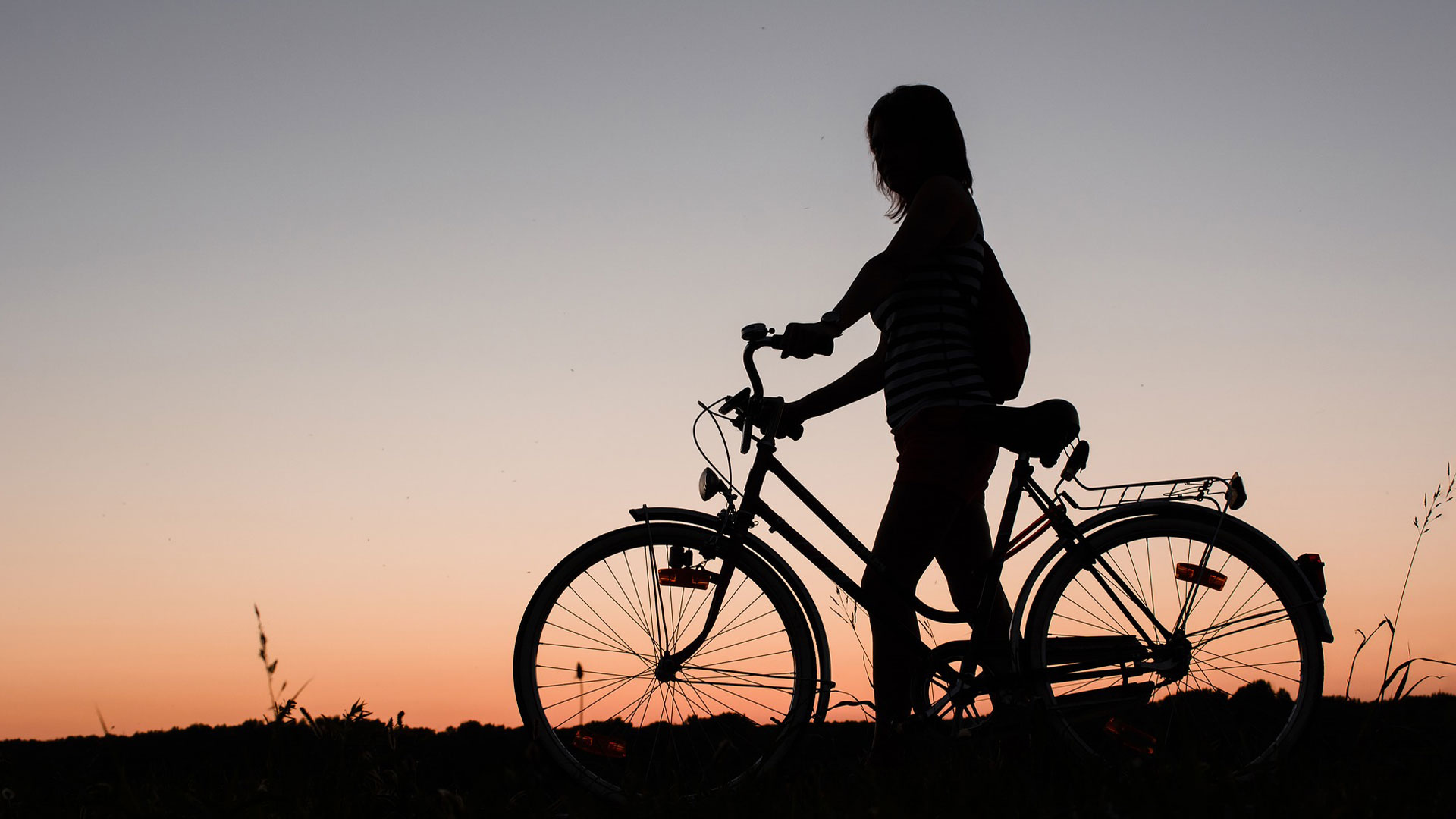 Siluett av en kvinna med cykel mot solnedgång.